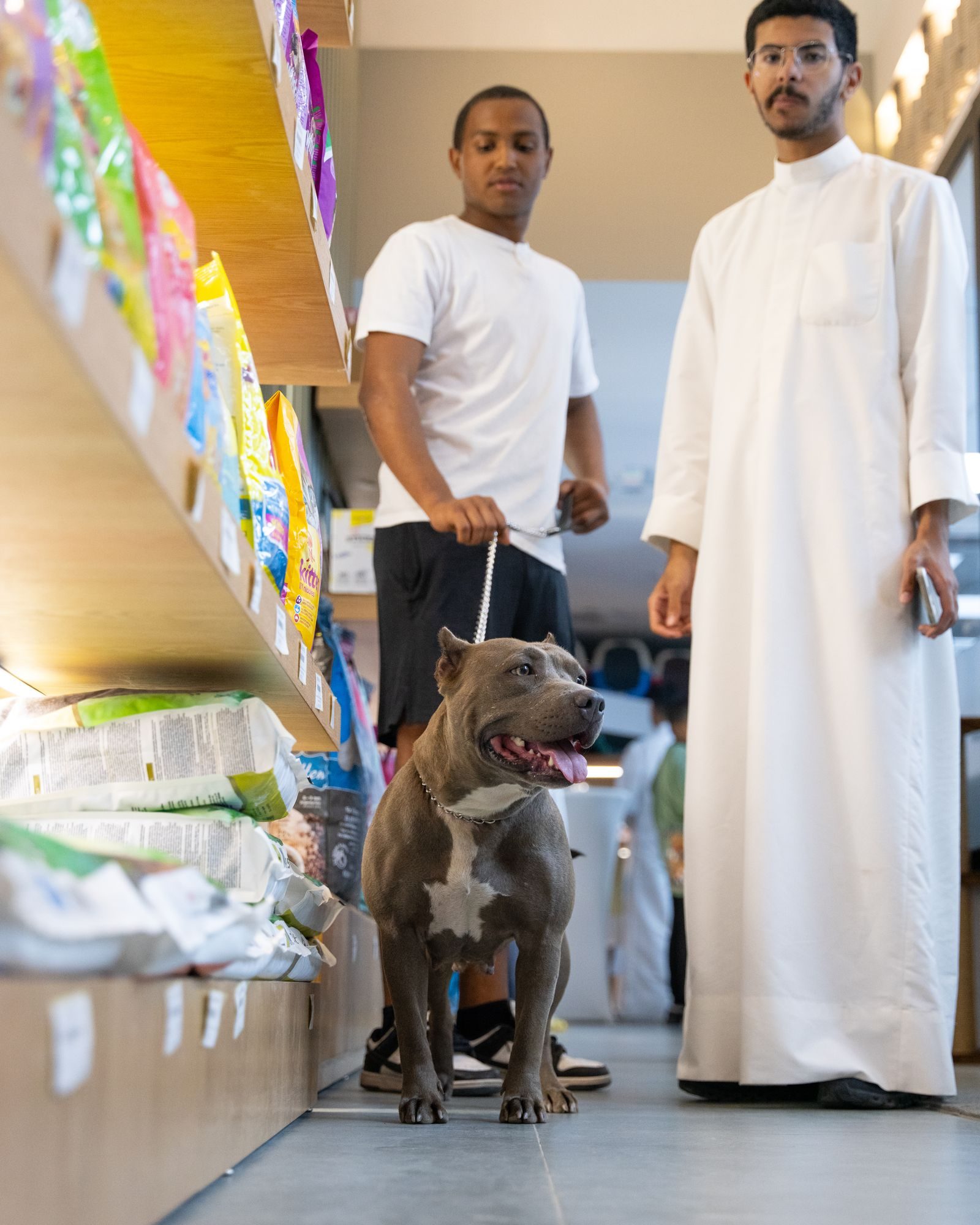 Two customers and a leashed dog beside shelves of pet food in a pet store.
