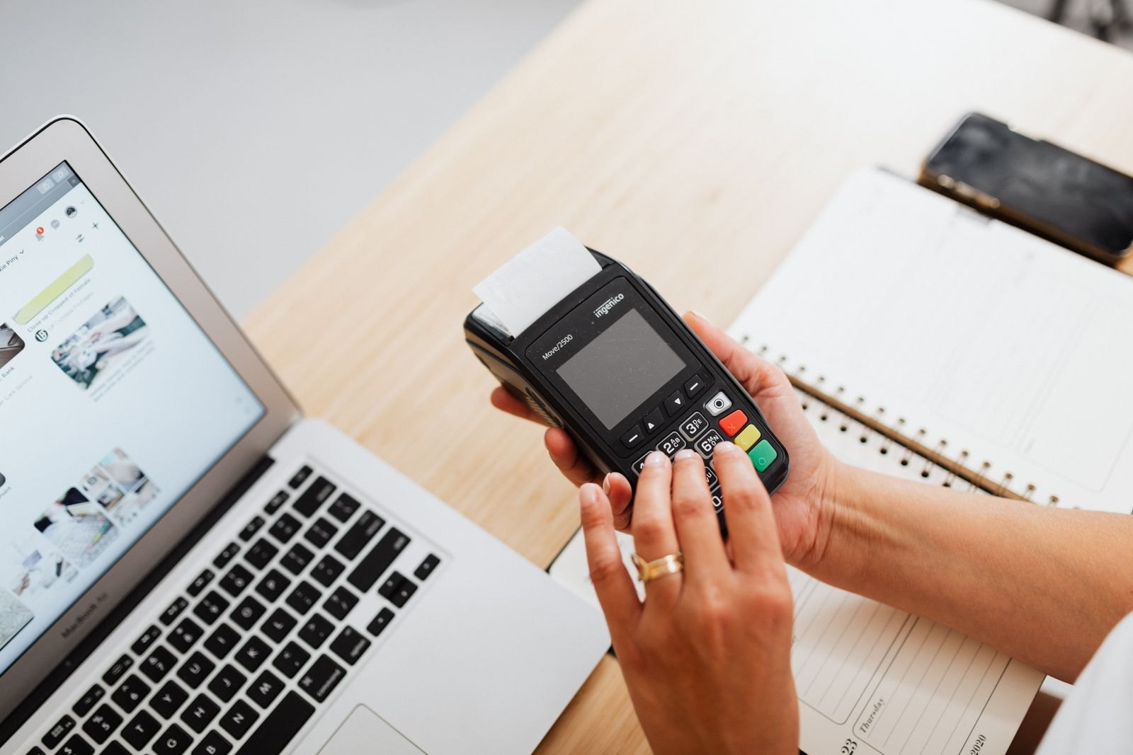 Hands operate a card terminal beside an open laptop and a notebook on a desk.